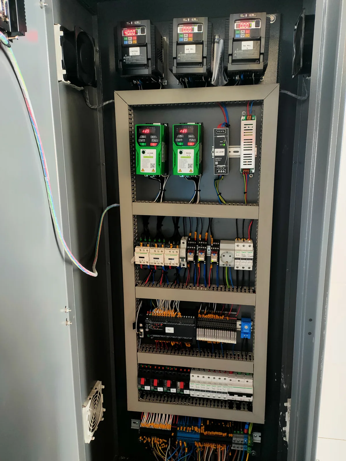 Open grey control cabinet with three drive units at the top and shelves of relays, breakers and terminal blocks below