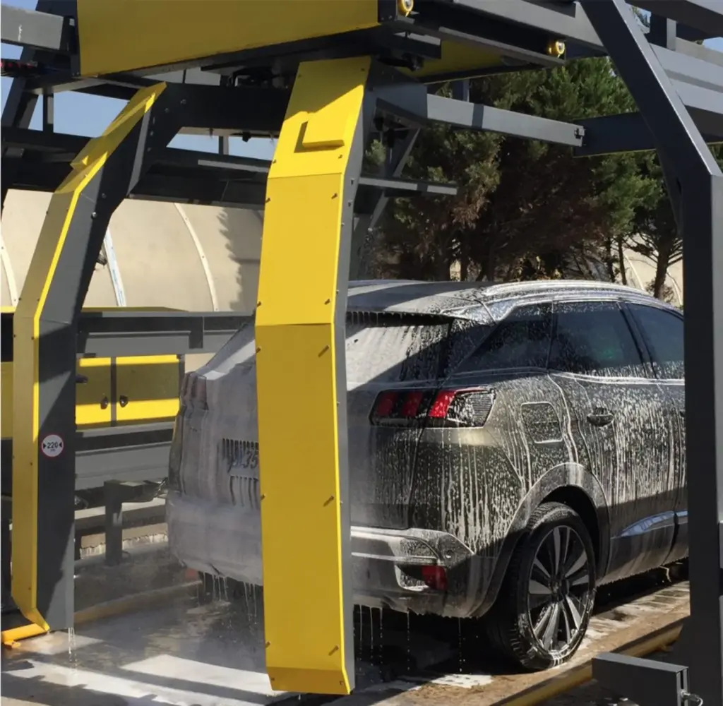 Dark SUV covered in white foam, standing under the grey and yellow arms of a rollover wash